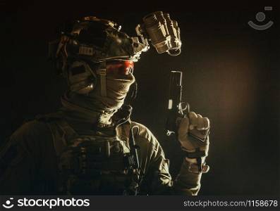 Side view portrait of army soldier, modern combatant, special forces fighter in helmet, night-vision device, radio headset, hiding identity behind mask, armed service pistol, low key studio shoot. Combatant armed with service pistol in darkness