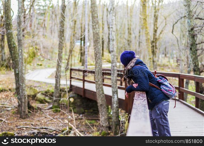 Side view portrait of beautiful girl with backpack standing near old footbridge. She is having break during hike in coniferous forest