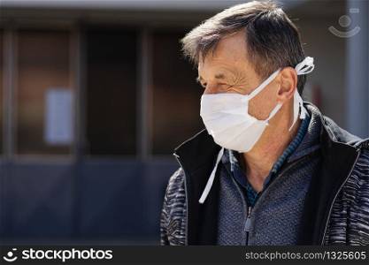 Side view portrait of caucasian senior man standing outdoor in front of the building or home wearing medical protective mask to prevent pandemic disease spread protecting from viruses pollution in day