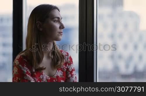 Side view portrait of thoughtful attractive female looking out of the window at home. Sad beautiful woman gazing through wide window. Pensive girl waiting for her boyfriend coming home and looking in window.