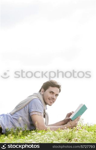 Side view portrait of young man holding book while lying on grass against clear sky