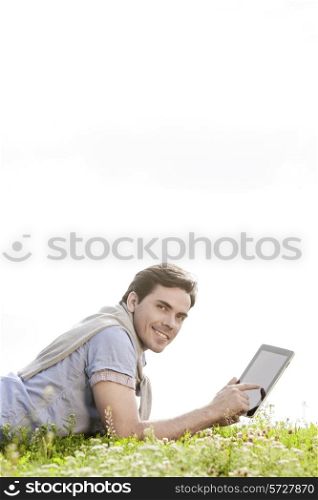 Side view portrait of young man using digital tablet while lying on grass against clear sky