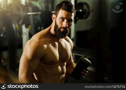 Side view portrait of young muscular caucasian man bodybuilder shirtless male sitting in dark gym holding protein supplement shaker in training waist up black hair and beard looking to the camera