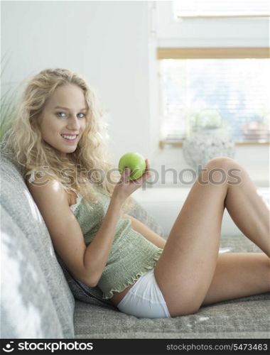 Side view portrait of young woman holding apple while sitting on sofa in house