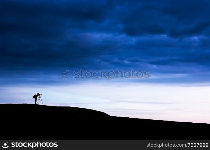 Side view shot of male photographer is taking images with dslr camera and tripod on the mountain peak under dramatic dark storm sky. Exploration concept.