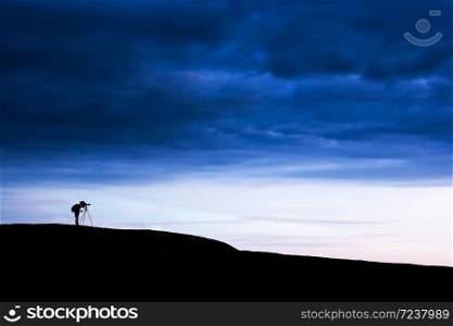Side view shot of male photographer is taking images with dslr camera and tripod on the mountain peak under dramatic dark storm sky. Exploration concept.