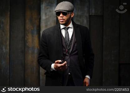 side view shot of stylish afro man in beautiful coat and glasses. Studio shot of stylish african american man wearing coat, cap, glasses and holding smoking pipe