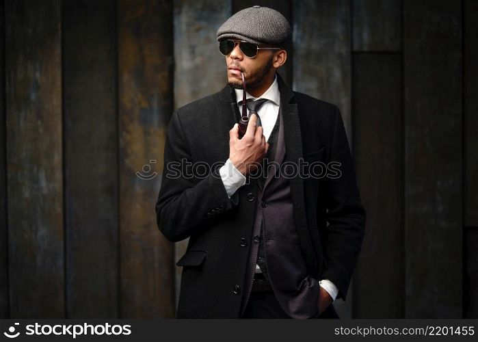side view shot of stylish afro man in beautiful coat and glasses. Studio shot of stylish african american man wearing coat, cap, glasses and holding smoking pipe