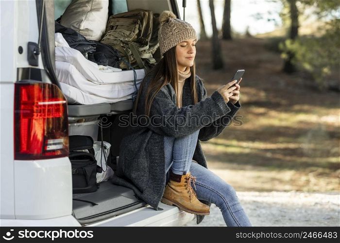 side view woman sitting trunk car while road trip using smartphone