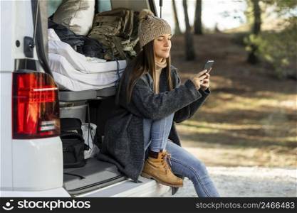 side view woman sitting trunk car while road trip using smartphone