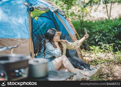 Side view Young Asian pretty woman and her girlfriend sitting at front of tent, use mobile phone take photo during c&ing in forest with happiness together