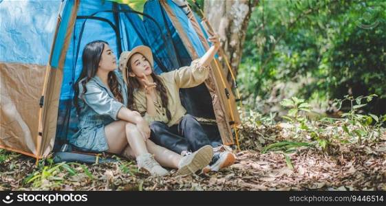 side view Young Asian pretty woman and her girlfriend sitting at front of tent, use mobile phone take photo during c&ing in forest with happiness together
