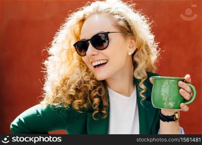 Sideways close-up of attractive woman wearing stylish shades and jacket holding green cup of tea having sincere smile on her face looking aside enjoying her life isolated over orange background