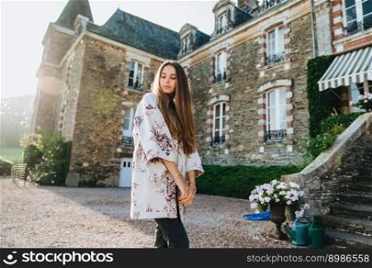 Sideways portrait of fashionable attractive young female with long hair, looks down with thoughtful expression, stands against big ancient builduing, thinks about something, poses outdoors