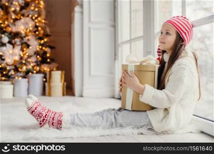 Sideways portrait of restful female child wears white sweater, trousers and warm socks, embraces wrapped gift, sits on floor in cozy room, admires New Year tree. Children and holidays concept