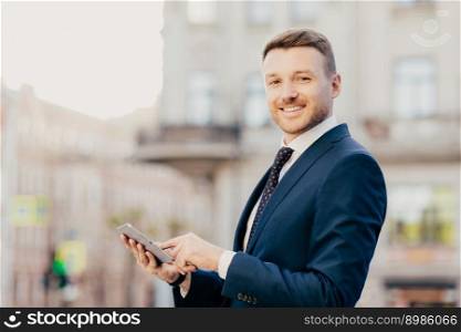 Sideways portrait of successful male freelancer works distantly on touchpad, stands outdoor, being in good mood after having dinner in luxury restaurant with colleague, connected to wireless internet