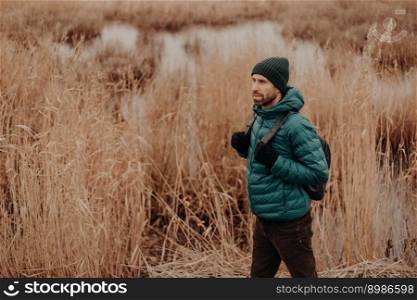 Sideways shot of active male dressed in warm jacket and hat, carries rucksack, has walk in rural area, poses near lake background, enjoys fresh air, calm atmosphere. People and lifestyle concept
