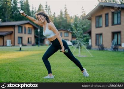 Sideways shot of active smiling determined woman raises dumbbells and has morning workout, dressed in active wear, poses on green lawn against private houses. Sportswoman trains biceps with weights