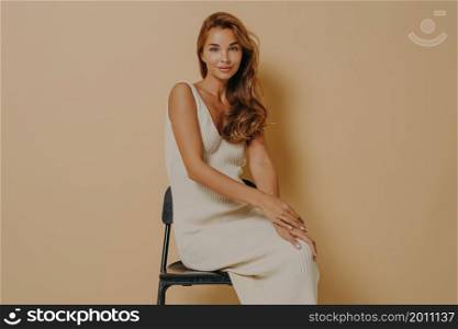Sideways shot of beautiful lady with wavy hair natural beauty and manicure wears long dress poses on chair against beige background has satisfied expression. Relaxed young European woman indoor. Sideways shot of beautiful lady with wavy hair poses on chair against beige background