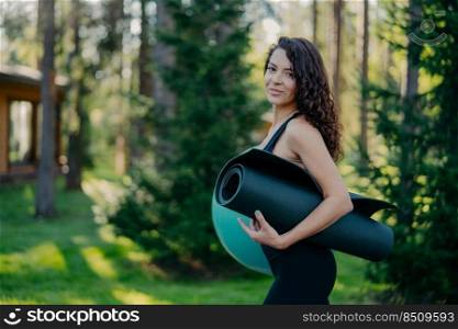 Sideways shot of beautiful slim woman with perfect figure, wavy dark hair, holds rolled up karemat and fitness ball, poses outdoor against blurred forest background, has aerobics exercises in morning