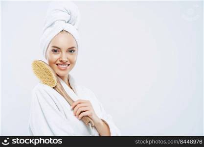 Sideways shot of beautiful young European woman with charming smile, poses with bath item, uses brush to massage body, poses against white background, blank space for your promotional content