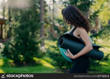 Sideways shot of brunette woman carries fit ball and rolled up karemat going to have fitness exercise turns away doesnt look at camera poses against blurred green nature background. Sporty lifestyle