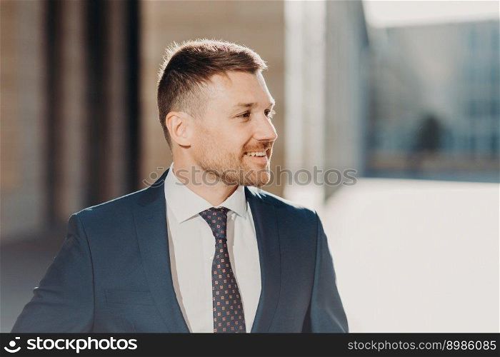 Sideways shot of cheerful unshaven male looks aside with joyful expression, wears formal clothes, being in good mood after successful meeting with business partners. People and work concept.