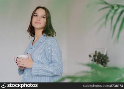 Sideways shot of dreamy Caucasian woman dressed in casual clothes, drinks tea, stands indoor against domestic interior with copy space for your advertising content. People, rest and drinking concept