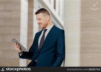 Sideways shot of elegant businessman in formal suit, holds digital tablet in hands and reads business news on internet website, poses in office, connected to wireless wifi. Modern technologies concept