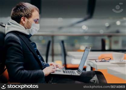 Sideways shot of freelancer keyboards on laptop computer, works on distance, wears protective medical mask during quarantine because of virus outbreak, poses in coffee shop, warns contagious disease