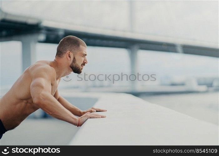 Sideways shot of handsome muscular man with thick beard does push ups exercise has workout outdoor leans on bridge fence engaged in physical activity poses with bare torso has strong muscles
