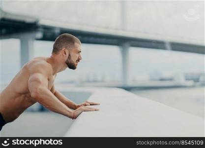 Sideways shot of handsome muscular man with thick beard does push ups exercise has workout outdoor leans on bridge fence engaged in physical activity poses with bare torso has strong muscles