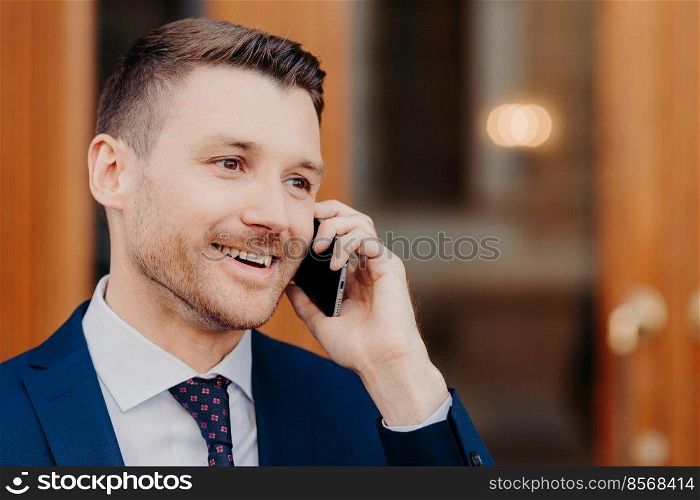 Sideways shot of handsome young male talks in roaming via cell phone, has positive expression, looks thoughtfully aside with copy space for business information. People and conversation concept