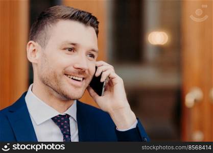 Sideways shot of handsome young male talks in roaming via cell phone, has positive expression, looks thoughtfully aside with copy space for business information. People and conversation concept