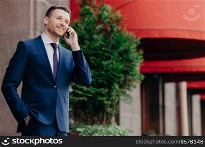 Sideways shot of intelligent male executive worker in formal suit, has telephone conversation via smart phone, keeps hand in pocket, enjoy tariffs for communication, stands outdoor in urban territory