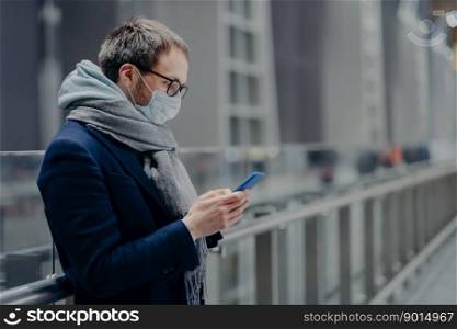 Sideways shot of male blogger sends text messages and reads comments from followers on modern cellphone, wears sterile medical mask to protect himself from coronavirus or other infectious disease