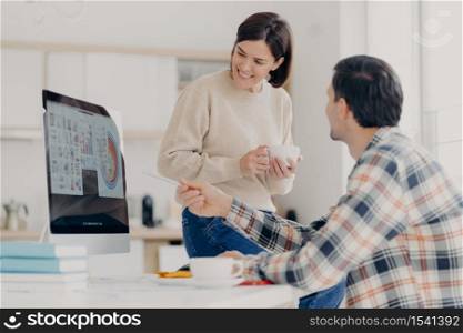Sideways shot of male financier tries to explain something to wife, points with pen at monitor of computer, prepares financial computer, plan to buy something expensive, collaborate together
