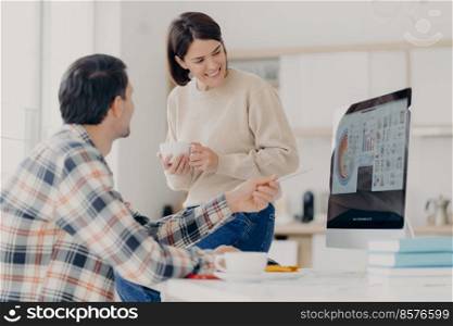 Sideways shot of male financier tries to explain something to wife, points with pen at monitor of computer, prepares financial computer, plan to buy something expensive, collaborate together