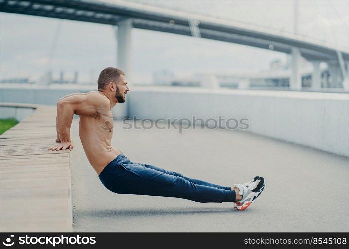 Sideways shot of muscular man does reverse push up exercise, trains arms, keeps perfect body and good physical shape, has active workout outdoor, wears sport trousers and sneakers, poses on bridge