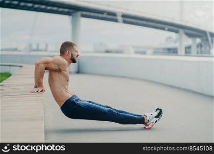 Sideways shot of muscular man does reverse push up exercise, trains arms, keeps perfect body and good physical shape, has active workout outdoor, wears sport trousers and sneakers, poses on bridge