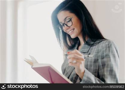 Sideways shot of pleasant looking brunette woman has happy expression makes notes in planner, does remote job, creats planning, wears optical eyeglasses and formal clothes, poses near window
