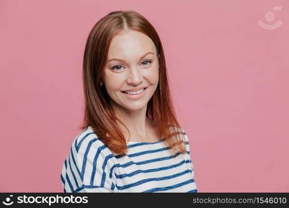 Sideways shot of pleasant looking happy brown haired woman with straight hair, healthy skin, pleasant smile, dressed in casual striped sweater, isolated over pink background. Emotions concept