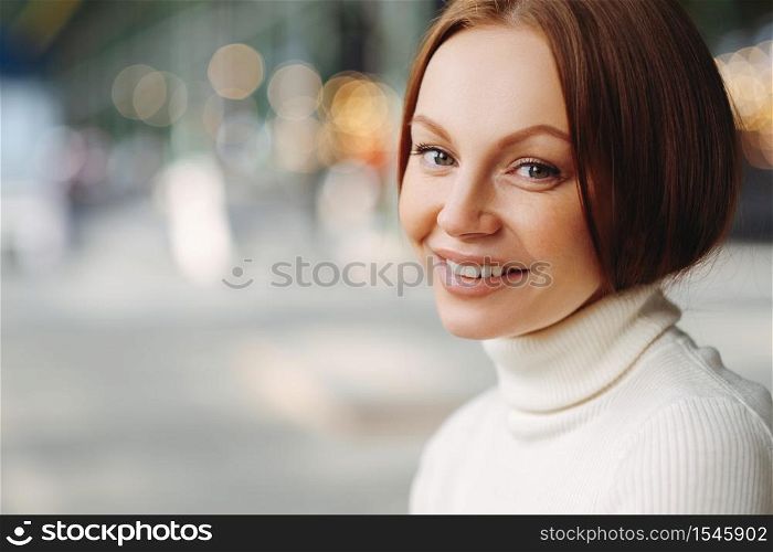 Sideways shot of pleasant looking woman with make up, tender smile, dressed in white turtleneck sweater, stands against blurred background with copy space for your advertisement, has healthy skin