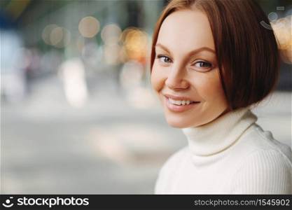 Sideways shot of pleasant looking woman with make up, tender smile, dressed in white turtleneck sweater, stands against blurred background with copy space for your advertisement, has healthy skin