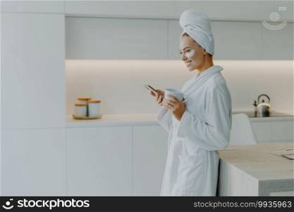 Sideways shot of pretty European woman feels refreshed after taking bath wears white soft bathrobe drinks hot beverage scrolls internet via mobile phone poses over kitchen interior wears beauty pads