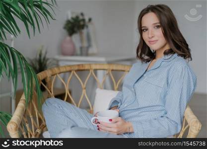 Sideways shot of pretty woman sits crossed legs in wicker chair, dressed in pyjamas, drinks hot coffee and latte, poses against cozy interior indoor, has weekend. People and relaxation concept