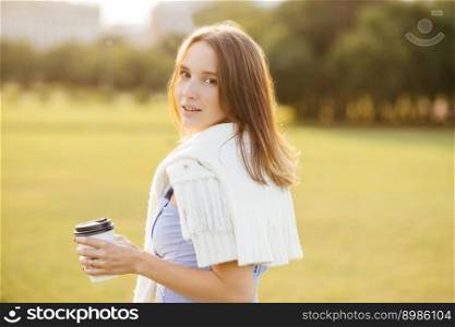 Sideways shot of pretty young girl with dark har, holds cup with coffee or cappuccino, breathes fresh air, enjoys calm atmosphere, admires beautiful nature and sunset. People and lifestyle concept
