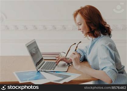 Sideways shot of redhead young Caucasian woman freelancer makes remote work, concentrated in laptop computer, sits at wooden desk with papers, takes off eyeglasses, surfs internet and reads news