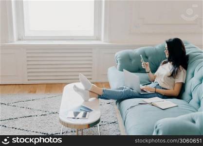 Sideways shot of relaxed female student uses modern technologies while doing homework, uses web sources for making project, sits on comfortable sofa against cozy interior, leans legs on desktop