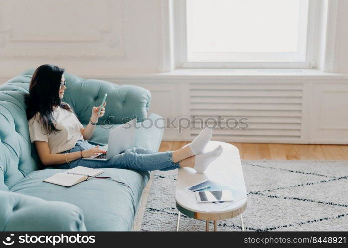 Sideways shot of relaxed female student uses modern technologies while doing homework, uses web sources for making project, sits on comfortable sofa against cozy interior, leans legs on desktop